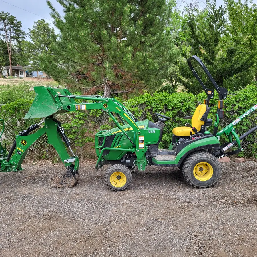 A green compact tractor with a front loader, a backhoe and a post-hole auger, parked on gravel in front of a hedge.